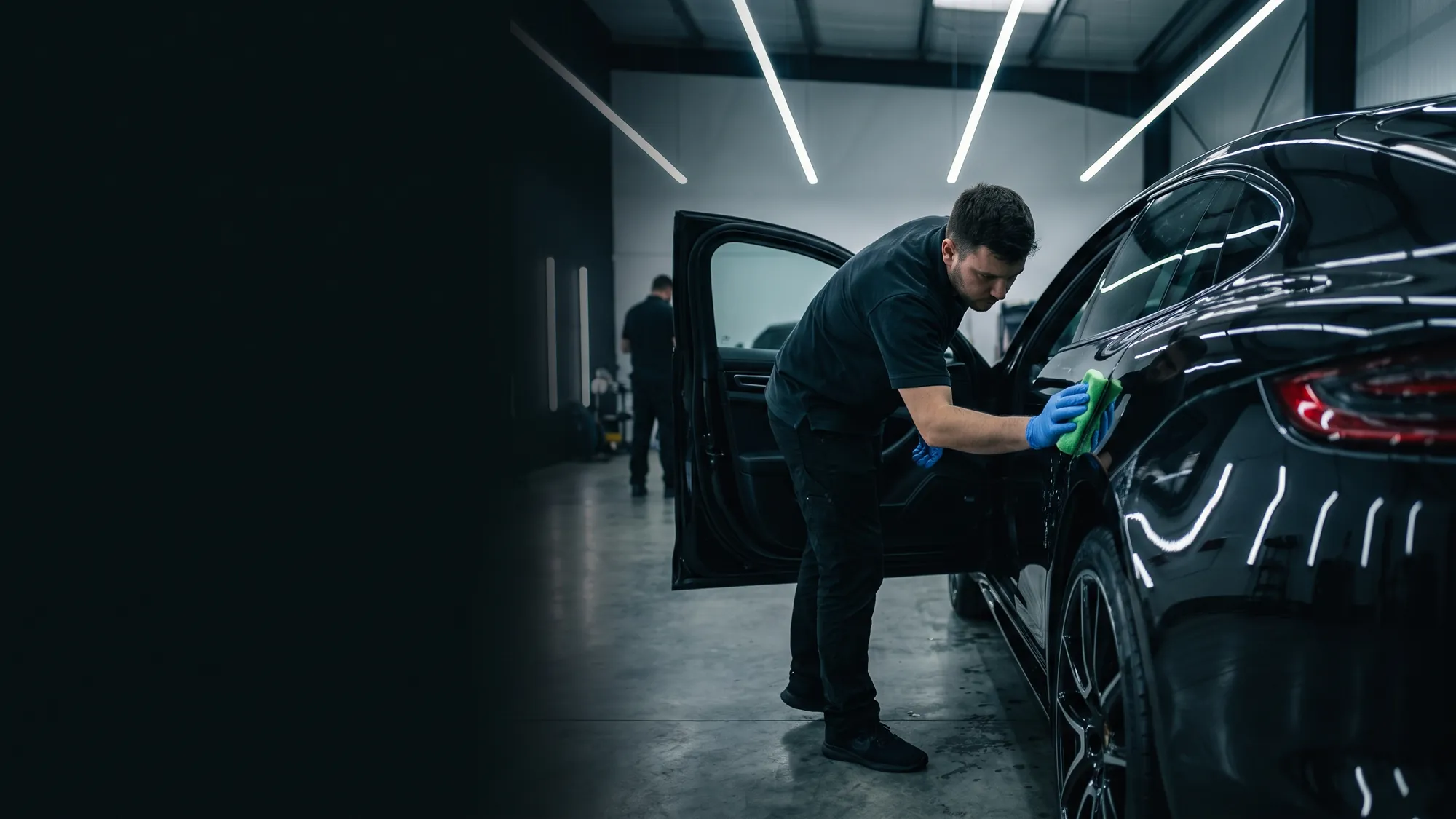 A technician in nitrile gloves applying ICON Rocklear to the door of a gloss black car with a microfibre applicator pad, in a certified bay