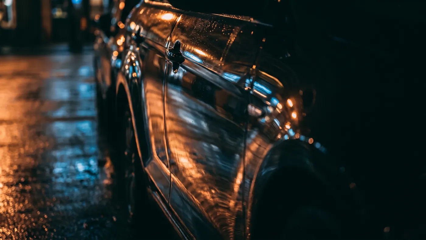 The flank of a black car at night mirroring city lights and a wet street