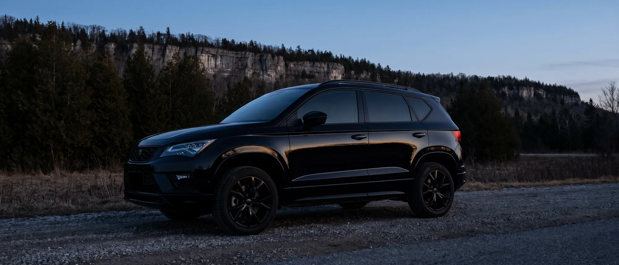 A gloss black SUV on a gravel shoulder below a limestone escarpment at last light