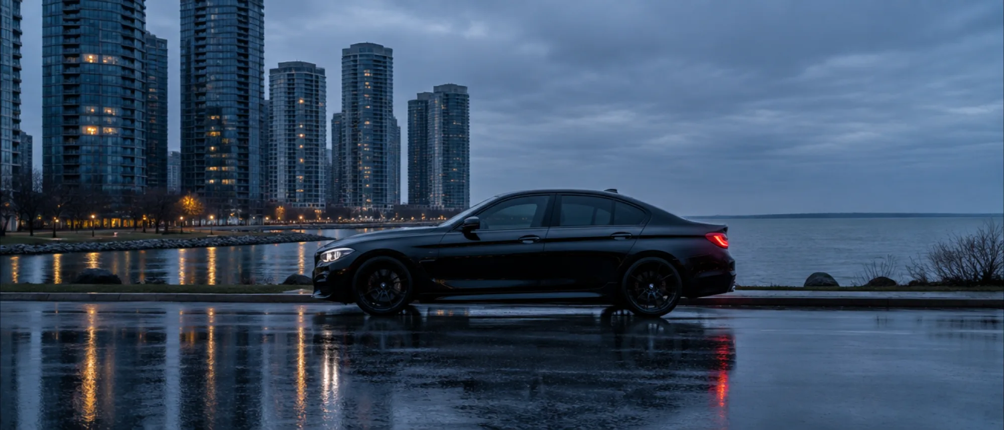A gloss black saloon in profile on a wet lakeside parkway at blue hour, glass towers and open water behind it