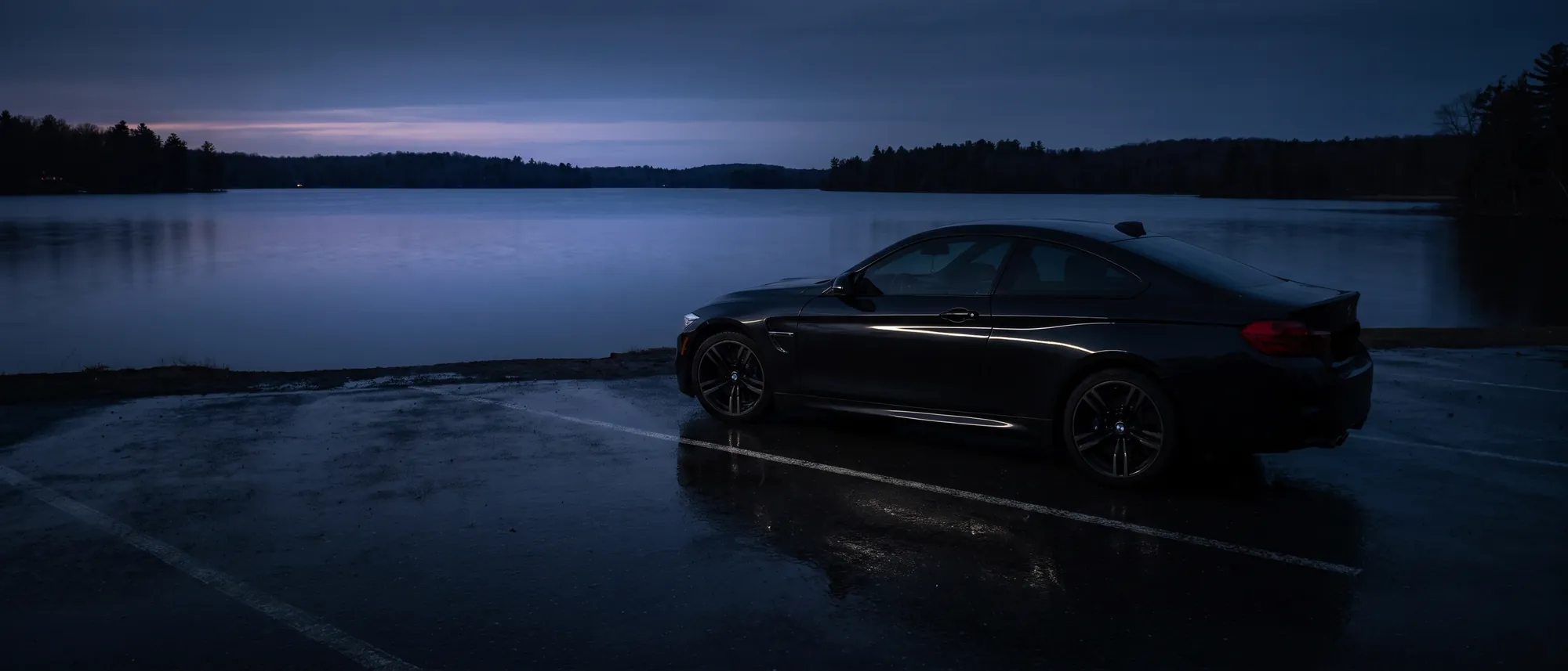 A dark coupe on wet asphalt beside still water at dusk, mirrored in the ground