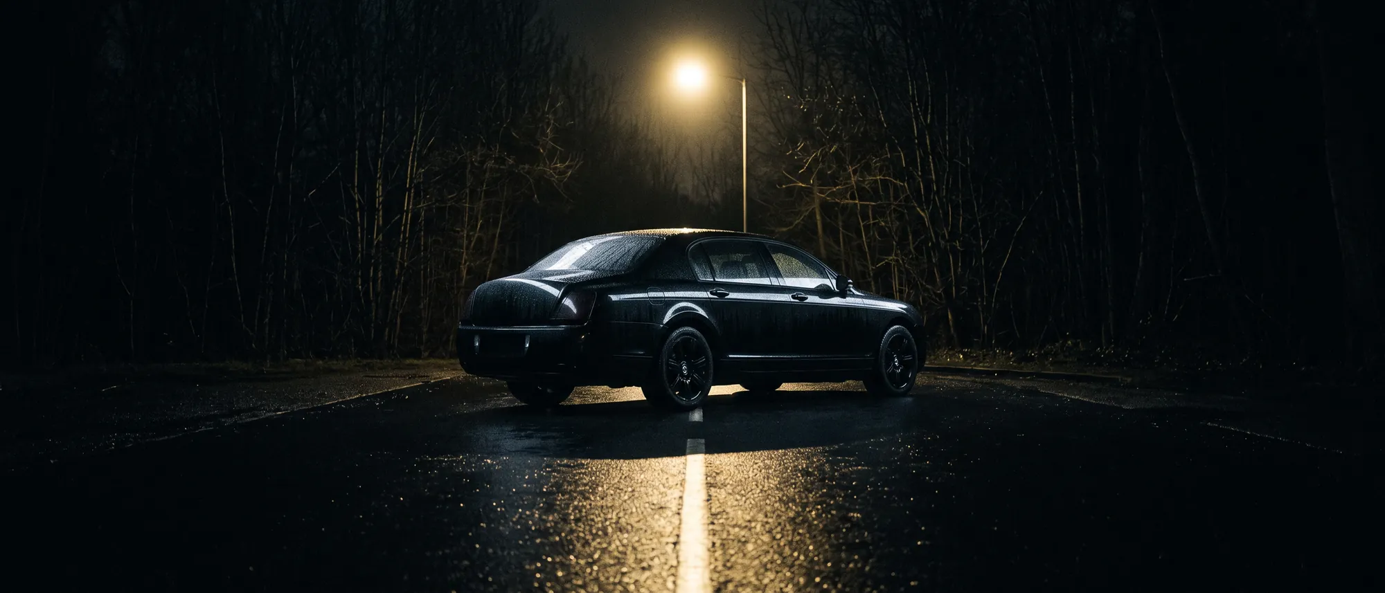 A black saloon on a wet road at night under a single street lamp