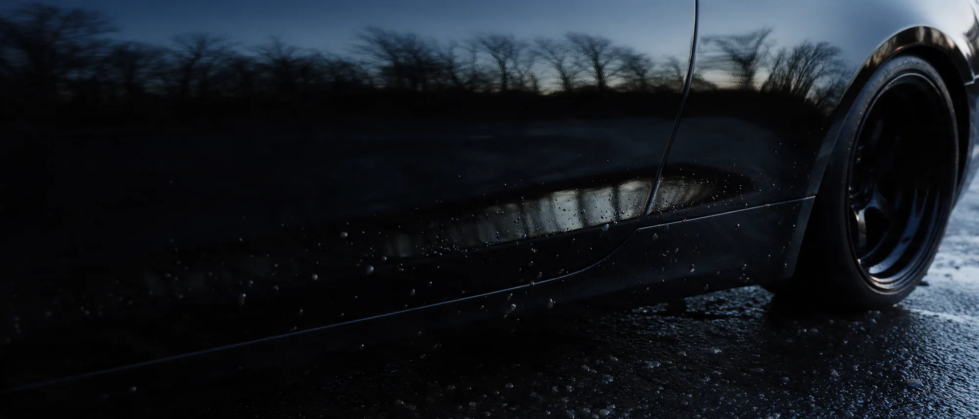 The coated flank of a dark car at dusk holding the reflection of a bare winter treeline