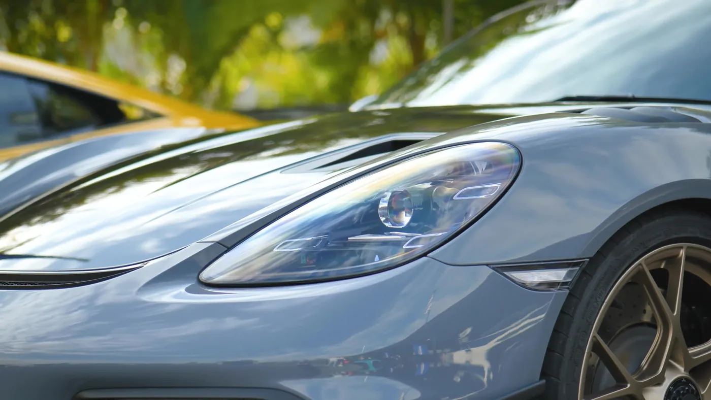 Macro of a coated Porsche 718 headlight and wheel with foliage reflected in the flank
